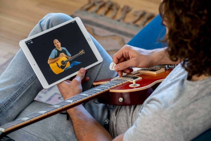 Person sitting down watching a guitar lesson on the tablet while holding a guitar pick with guitar on lap